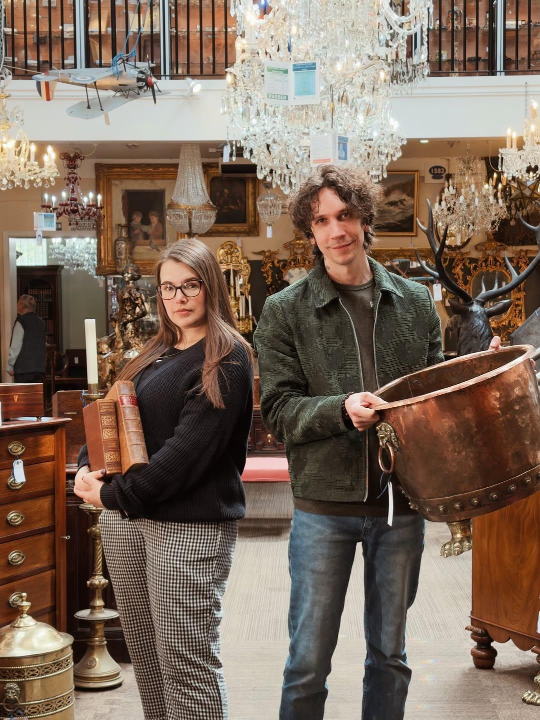 Cherry and Leslie are stood facing the camera in the middle of Hemswell Antique Centre. Cherry is holding two old leather books, whilst Leslie is holding a big copper cauldron. There are elaborate chandeliers above, and an entire room filled with antiques and artifacts all around them.
