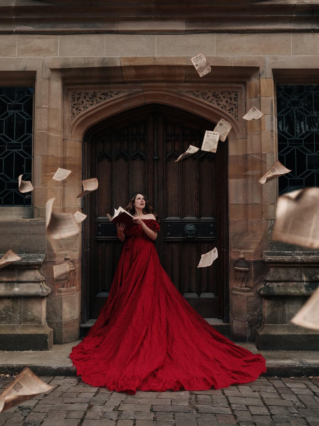 Cherry is stood in front of a grand wooden double door at Thornbridge Estate, with beautiful brick surrounding it. She’s wearing a bright red gown with a long train, holding a vintage book. There are individual fantasy pages flying around the scene.