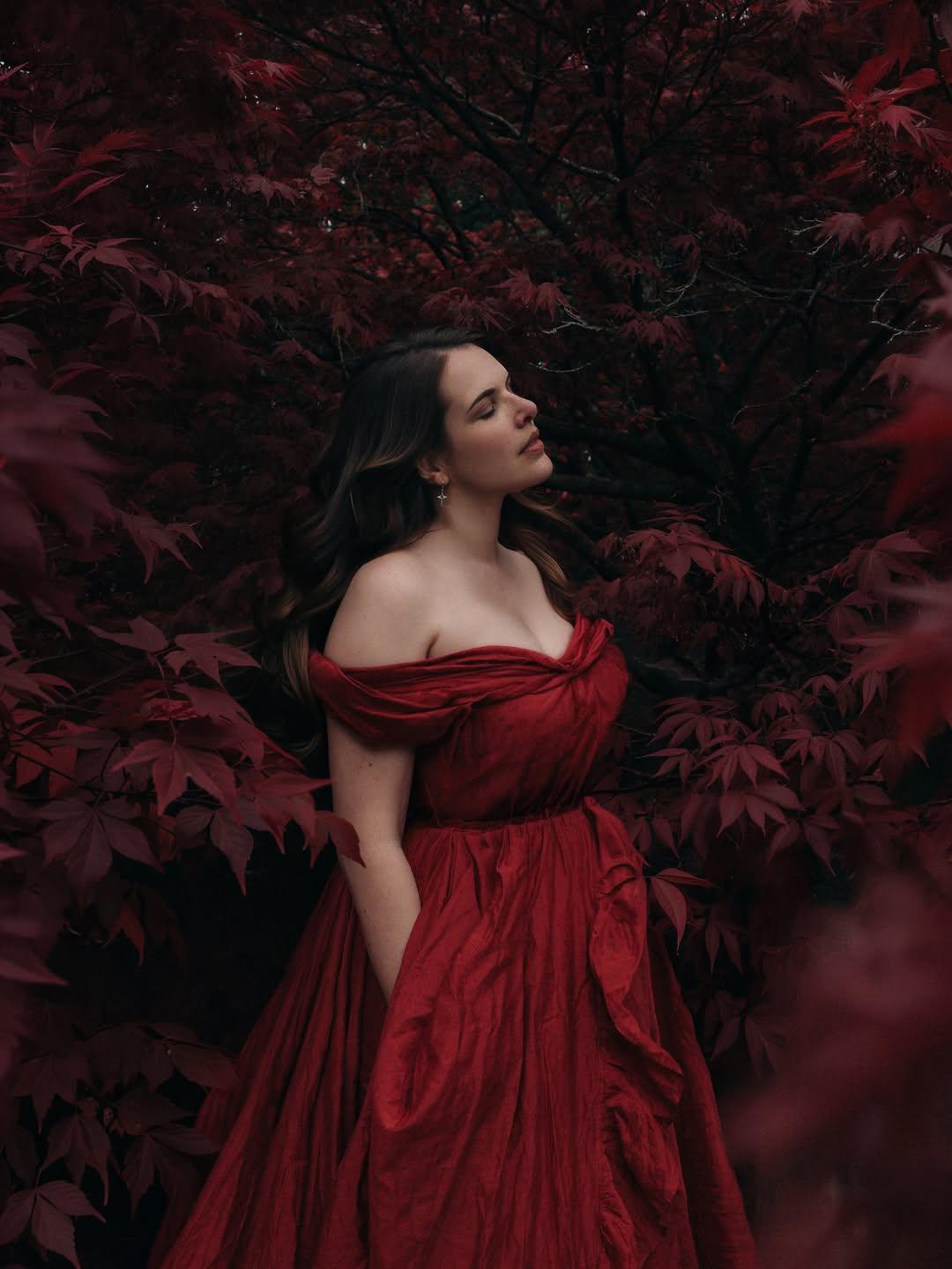Cherry is elegantly stood in a bright red ball gown, in front of an oriental type of tree, surrounded by deep autumn red leaves. She’s stood almost side on, with her eyes closed and face toward the sky.