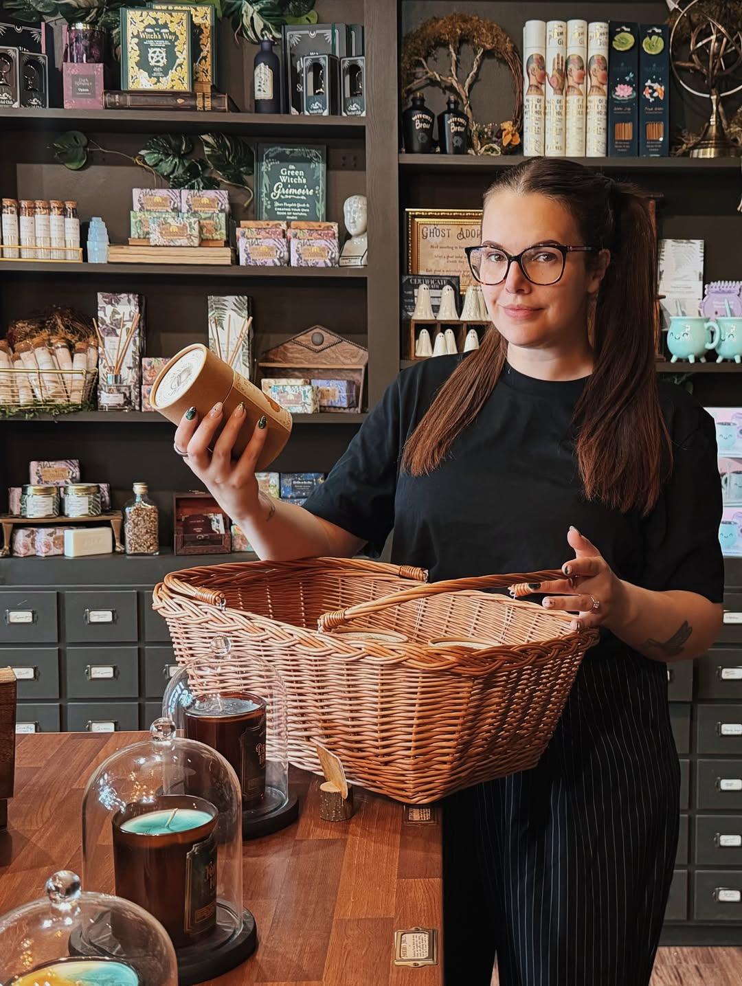 Cherry Wallis (co-owner of the lost cauldron shop) is inside her little shop, in work uniform, stood at a counter with a wicker basket full of stock to put out onto the shelves.