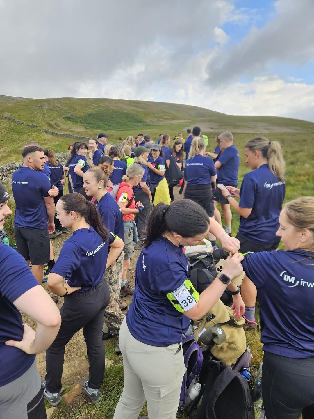 A group of people wearing matching navy blue shirts with "IM Group" printed on them, gathered on a grassy hillside. Some individuals are preparing for an outdoor activity, adjusting gear and talking. The background features green rolling hills and a partly cloudy sky.