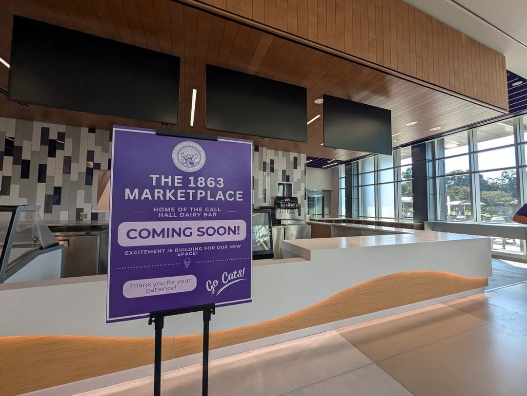 An empty marketplace area with modern decor and a wooden ceiling. In the foreground, there is a sign that reads "The 1863 Marketplace, Home of the Call Hall Dairy Bar, Coming Soon! Excitement is building for our new space! Thank you for your patience!"