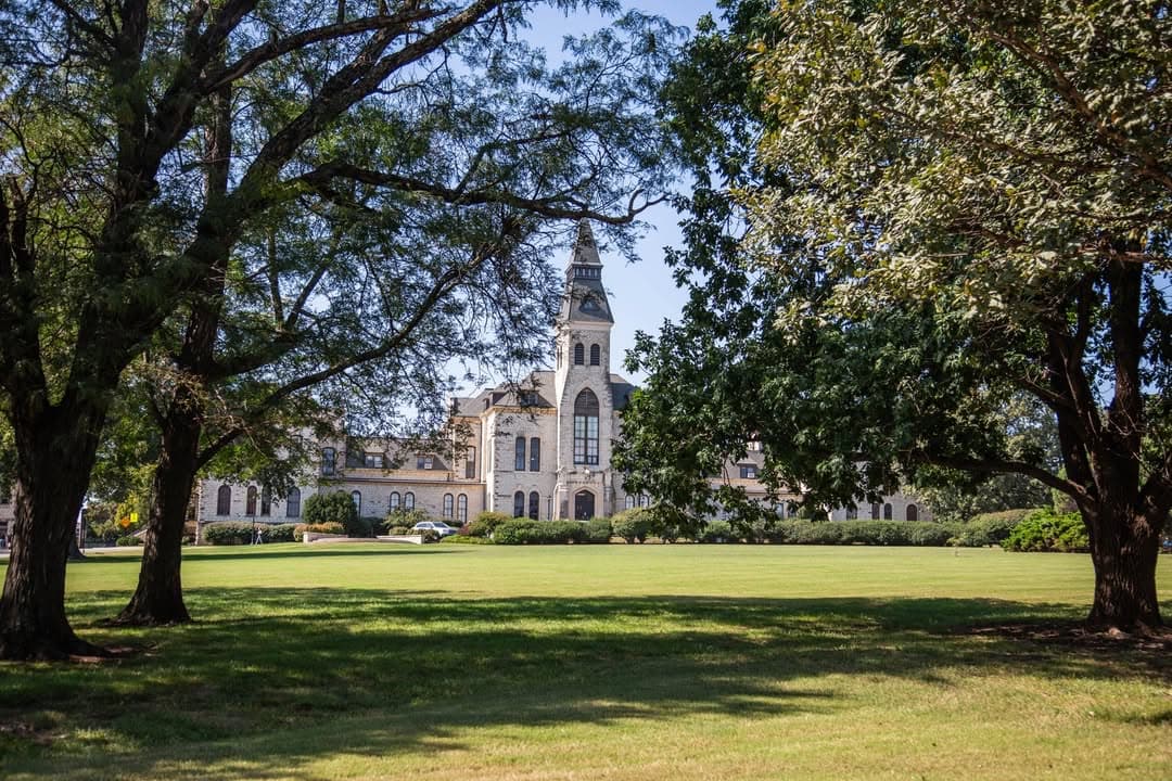 Anderson Hall is visible through a frame of trees, set against a clear blue sky. The scene includes a spacious, sunlit lawn in the foreground.