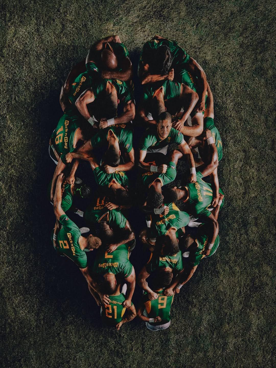Overhead view of the South Africa Springboks Rugby Team gathered tightly in a circular scrum. Players wear matching green jerseys with gold accents, forming a powerful, unified shape against the dark, textured grass. One player near the center looks upward while teammates link arms and lean together. The dramatic lighting creates strong contrast and highlights the team's formation which resembles a brain.