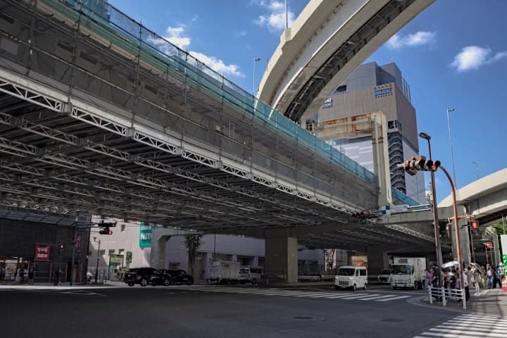 東京・池袋のサンシャイン前交差点付近。首都高速5号池袋線の高架下に、システム吊足場「クイックデッキライト」が設置されている。銀色の格子状の骨組みと床パネルで構成された作業床が、高架に沿って道路の上に広がっている。